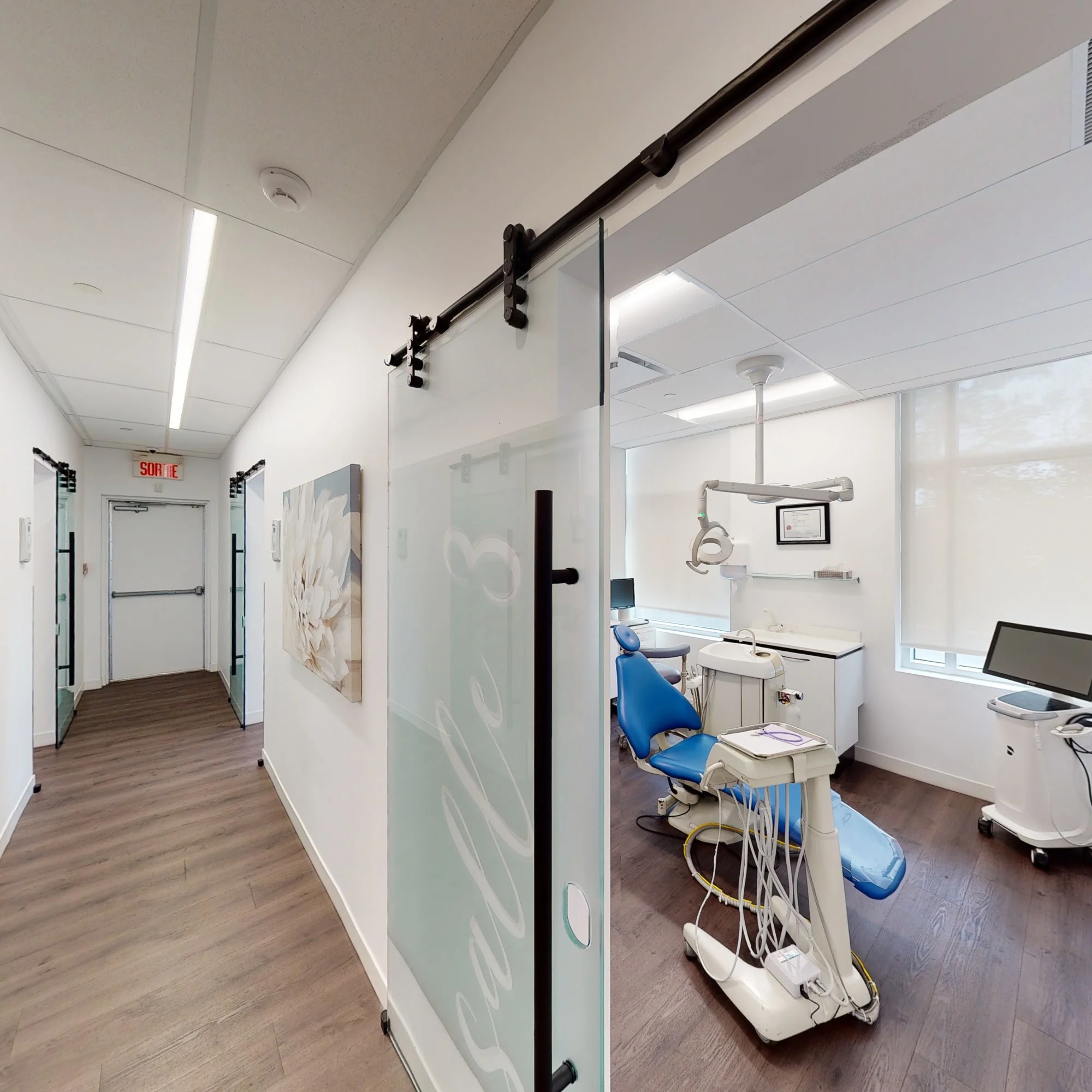 Couloir de la clinique ouvert sur une salle de soins au fauteuil bleu.