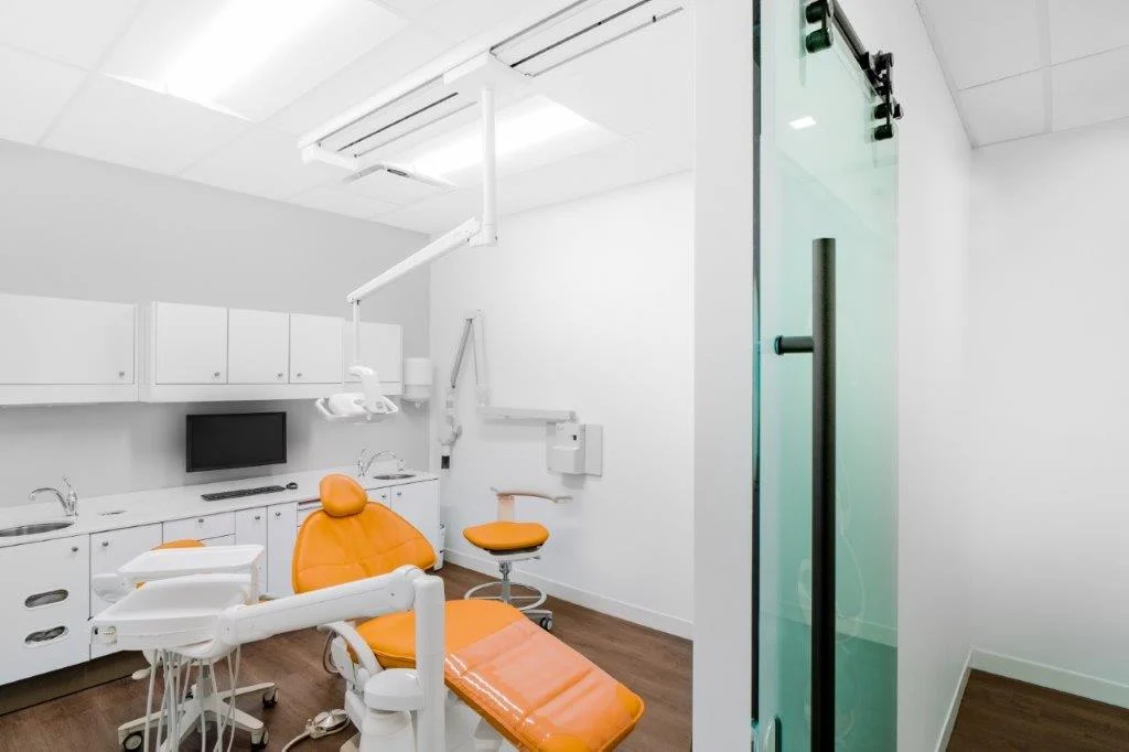 Salle de soins lumineuse avec fauteuil orange, vue depuis la porte vitrée.